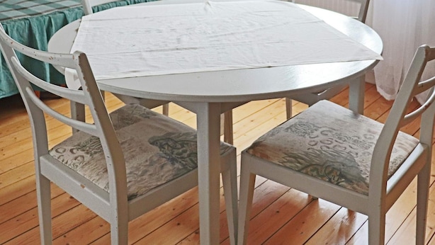 A round white table with four chairs, each with a patterned cushion, in a room with wooden flooring and a bed with a green checkered bedspread. - 9 Person Holiday Home in Bastad (Bastad)