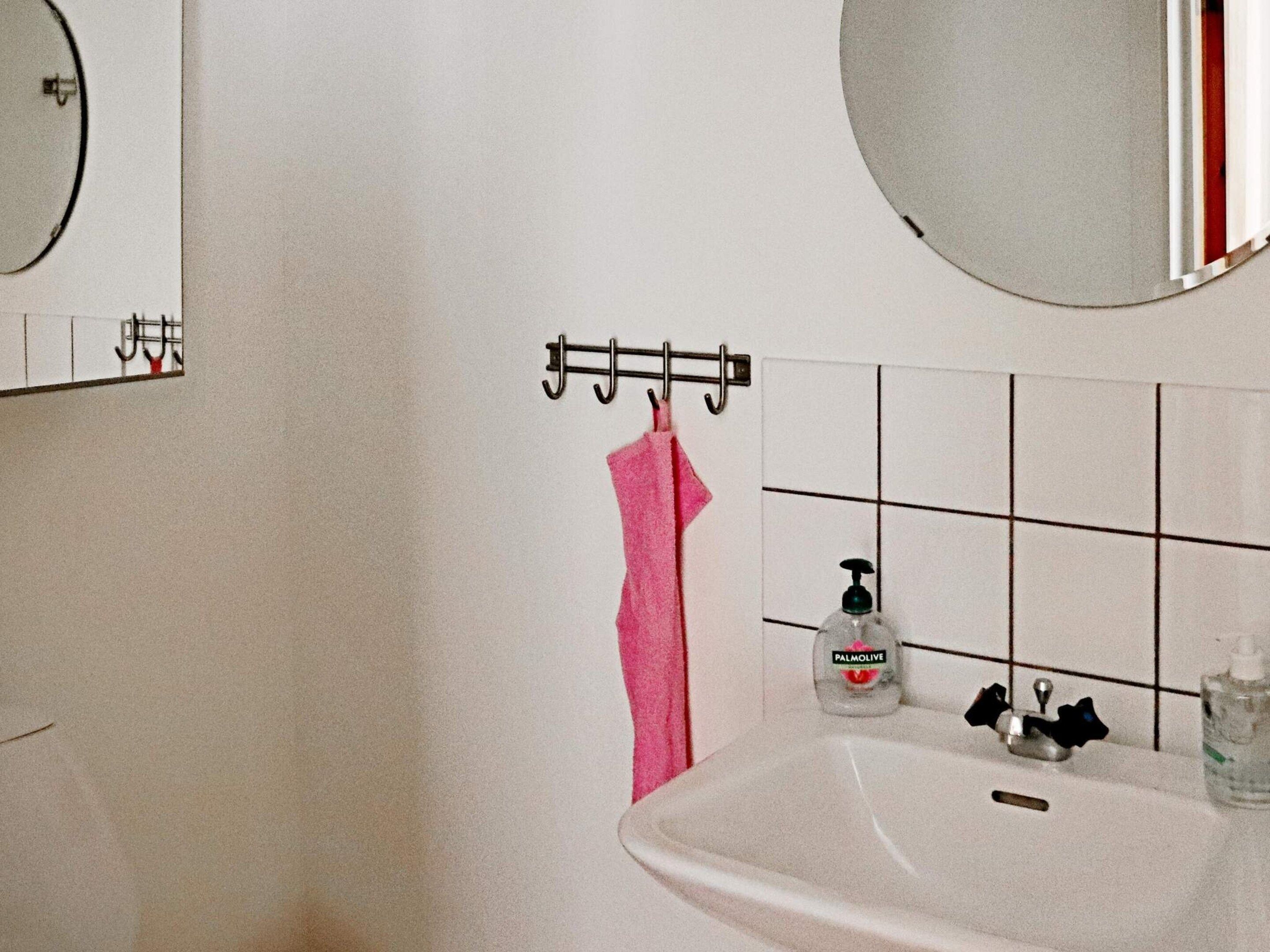 Et badeværelse med en hvid vask, et spejl, en håndklæderække med et pink håndklæde og to flasker sæbe.