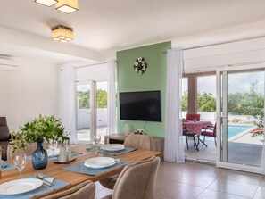 A dining area with a table set for two, a television mounted on the wall, and a view of a swimming pool through glass doors. - Villa Lena Comfortable Holiday Residence (Marcana)
