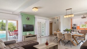 A modern living room with a large sofa, a coffee table, and a flat-screen TV. There is a dining area with chairs and a table, and a kitchen area with a microwave and cabinets. - Villa Lena Comfortable Holiday Residence (Marcana)