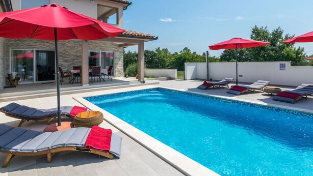 Une piscine avec des chaises longues et des parasols rouges, un patio couvert et un bâtiment en pierre doté de portes vitrées.