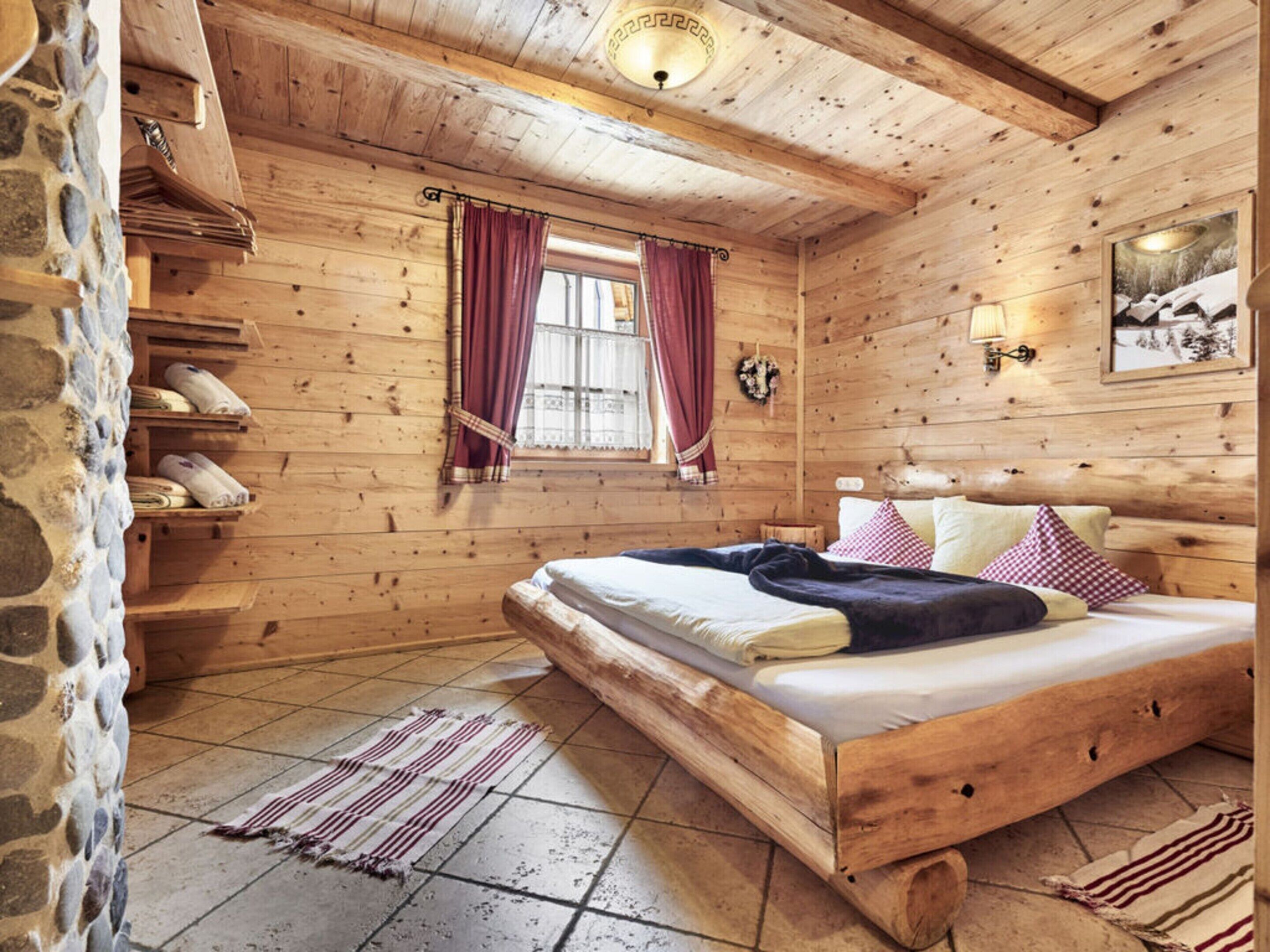 Ein Schlafzimmer einer Holzhütte mit Bett, Steinwand, Holzregalen, einem Fenster mit Vorhängen und einer Wandleuchte.