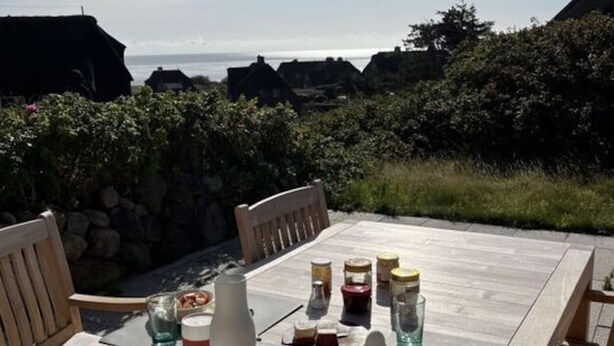 A wooden table set outdoors with various condiments and a bowl of food, overlooking a coastal area with houses and greenery. - Klaar Kimming Modern Retreat (Sylt)
