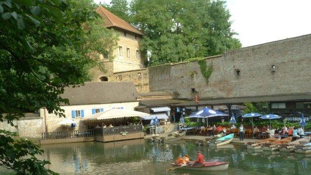 A riverside area with boats, outdoor seating, and a historic building. - Holiday Apartment Zirbelnuss (Augsburg)