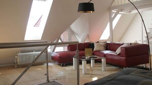 A high-ceilinged room with a red leather sofa, a glass coffee table, and a black floor lamp. A white radiator is visible against the wall, and a staircase leads to an upper level. - Holiday Apartment Zirbelnuss (Augsburg)