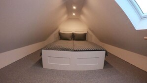 Una camera da letto mansardata, piccola e con tetto spiovente, presenta un letto al centro sotto una finestra a lucernario, con una semplice testiera bianca e due cuscini.