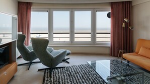A modern hotel room with a large window offering a view of the beach, a grey armchair, an orange sofa, and a glass coffee table.