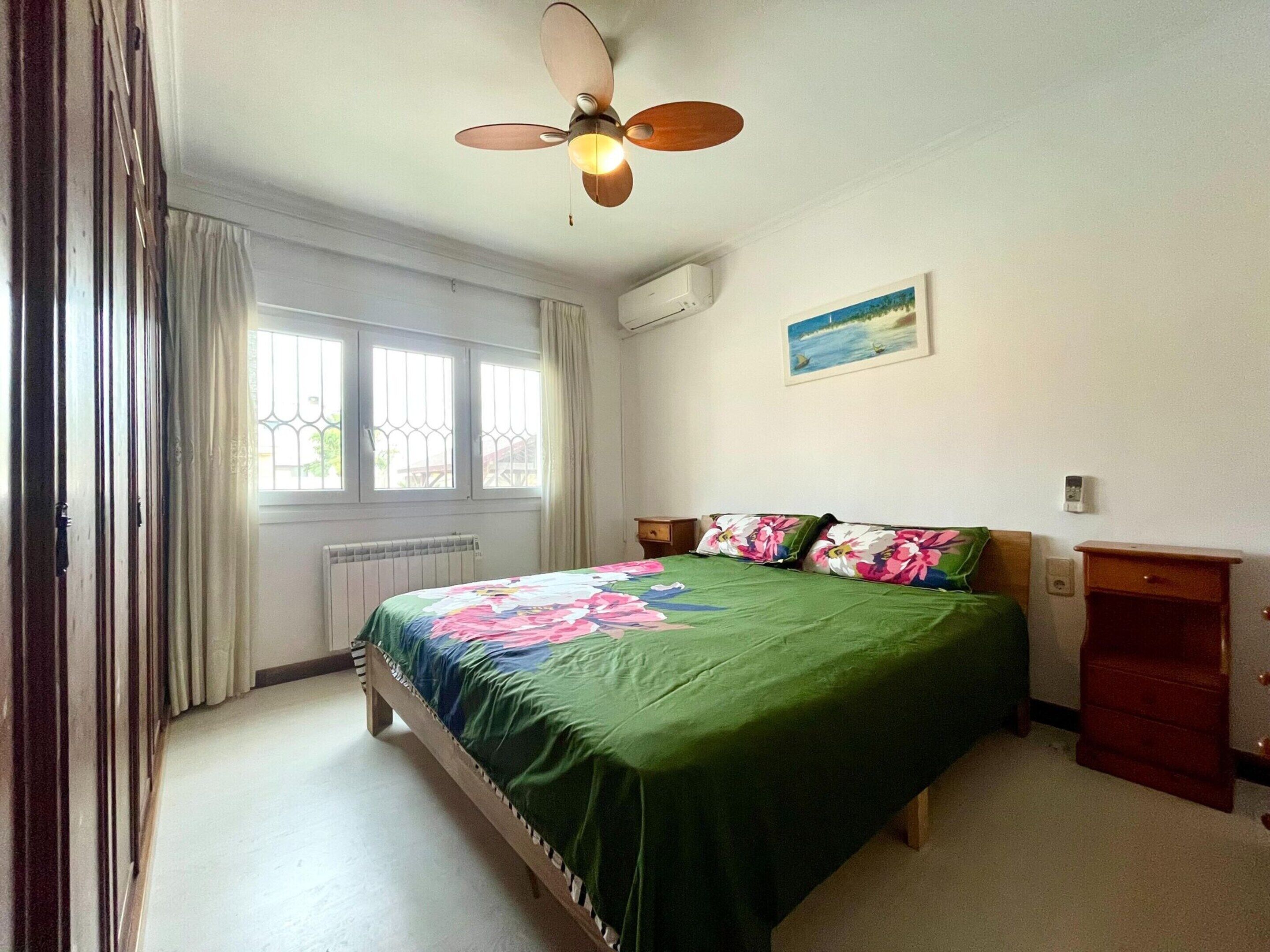 A bedroom with a bed, a ceiling fan, a window, a radiator, and a nightstand.