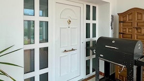 Une porte blanche avec une poignée en laiton et une grille noire, un meuble en bois sur la droite, et une lanterne fixée au mur au-dessus.