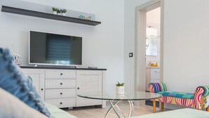 Una sala de estar moderna con un televisor de pantalla plana, muebles blancos, una mesa de centro de vidrio y un sofá colorido.
