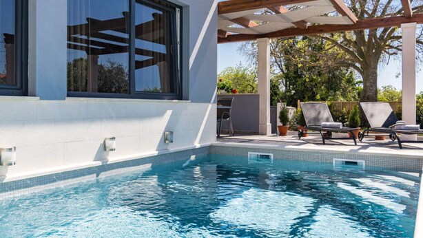 Un área de piscina moderna al aire libre con una pérgola, sillones de descanso y un amplio ventanal con vistas a la piscina.