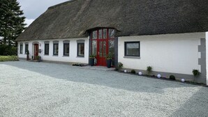 Uma casa de campo tradicional com telhado de colmo, porta vermelha e paredes brancas, entrada de cascalho e plantas em vasos ao longo do lado.