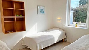 A small, cozy room with a single bed, a wooden shelf unit, and a window with a view of greenery.