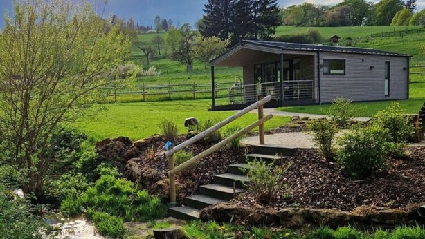 Un edificio moderno con un espacio exterior cubierto, rodeado de un paisaje verde exuberante y un pequeño arroyo.