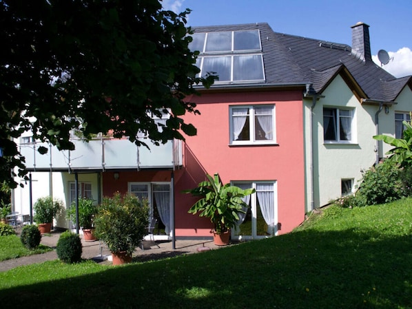 A two-story house with a red facade, white windows, and a grey roof. There are potted plants and a grassy lawn in front.