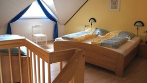 A bedroom with a sloped ceiling, a wooden bed, a chair, and a window with blue curtains. - Muhlenhof - Muller Thurgau (Maring-Noviand)