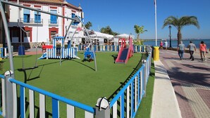 A playground with swings and slides, a fenced area, and a building with red and white walls in the background. - Belvilla by OYO La Tercia Resort (Murcia)