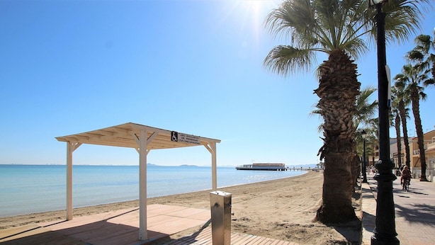 A beach with a wooden pier, a covered seating area, and a palm tree. - Belvilla by OYO La Tercia Resort (Murcia)