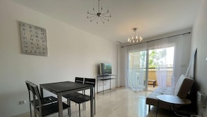 A modern living room with a dining area, a sofa, a TV, and a large window with curtains. - Belvilla by OYO La Tercia Resort (Murcia)