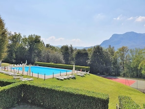 A swimming pool with lounge chairs, a tennis court, and a mountainous backdrop. - Apartment Near Lago Maggiore and Lugano (Montegrino Valtravaglia)