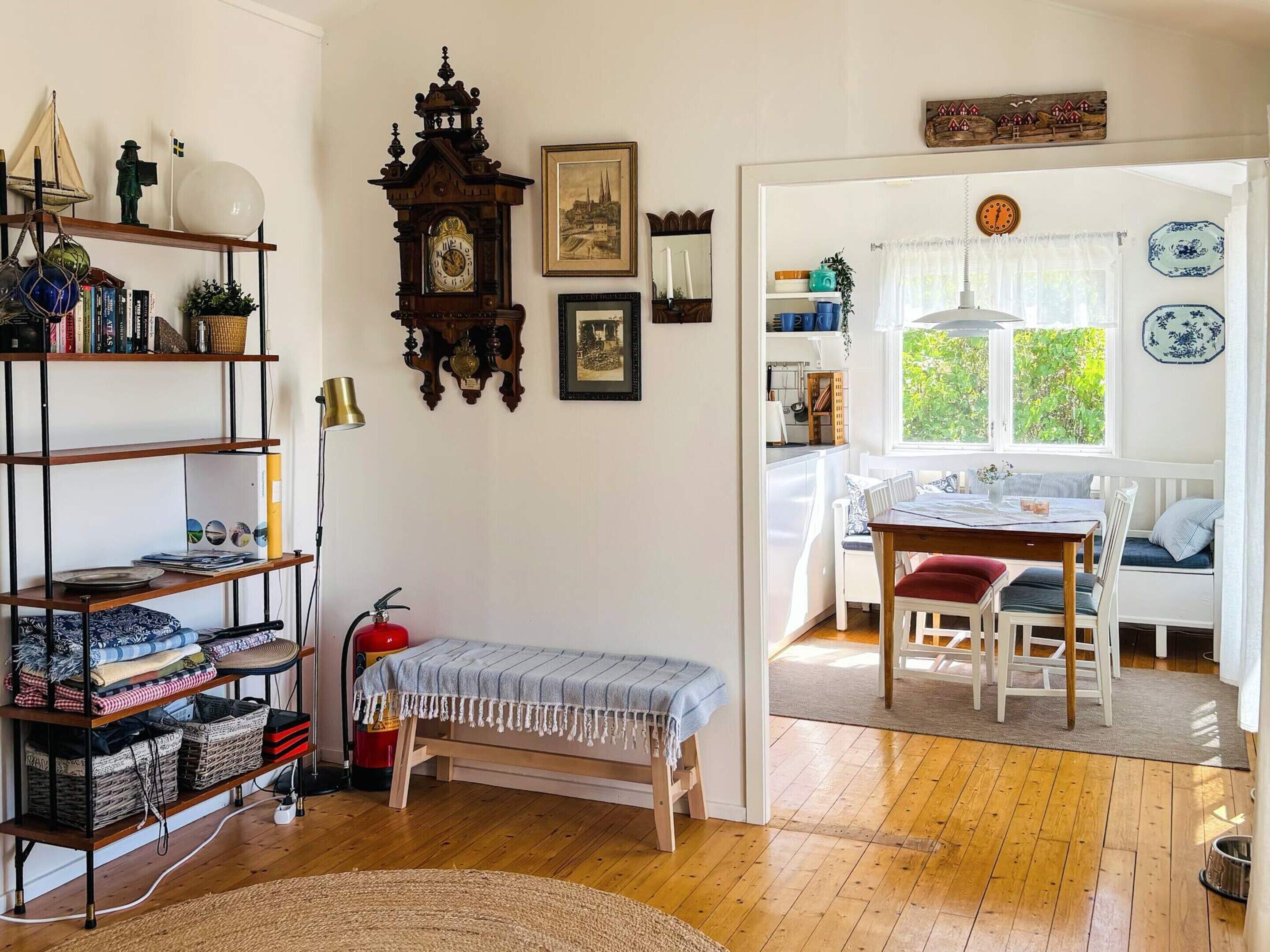 Una habitación con suelo de madera, un banco, una estantería, un reloj de pared y una zona de comedor con mesa y sillas.