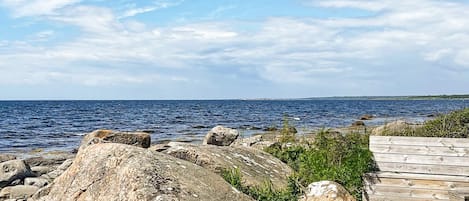 Ett kustlandskap med en träbrygga som vetter mot havet, omgivet av stora stenar och frodig grönska.
