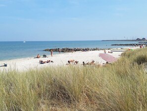 Une plage où des gens se prélassent au soleil et se baignent, une jetée en arrière-plan et un espace herbeux au premier plan.