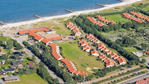 Luchtbeeld van een kustresort met meerdere bungalowachtige gebouwen, een zandstrand en een rotsenrijke zeewering.