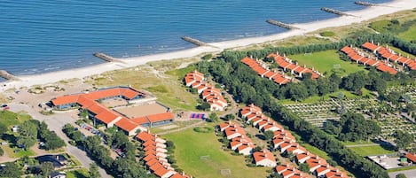 An aerial view of a coastal resort with multiple bungalow-style buildings, a sandy beach, and a rocky breakwater.