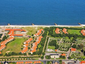 Vue aérienne d’un quartier résidentiel situé sur la côte, avec des rangées de maisons, une plage et un grand bâtiment doté d’un toit rouge.