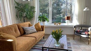 Un salón con un sofá marrón, un tapete a rayas, una mesa de centro con un florero y una ventana con plantas y una lámpara.