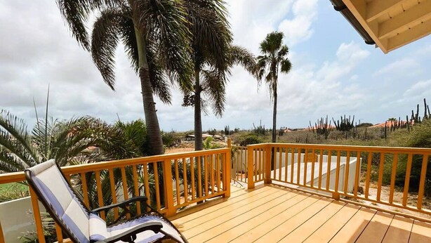 A wooden deck with a yellow railing, a lounge chair, and palm trees in the background. - Paradera Palms Apt. 2 (Paradera)