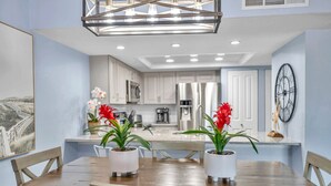 A dining area with a wooden table and chairs, potted plants, and a modern kitchen in the background. - Charming First Floor Condo at the Beach (Indian Shores)