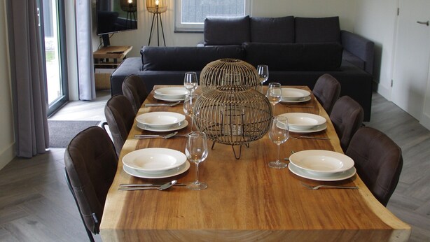 Un coin repas avec une table en bois dressée pour six personnes, entouré de canapés sombres et d’un téléviseur posé sur un meuble.