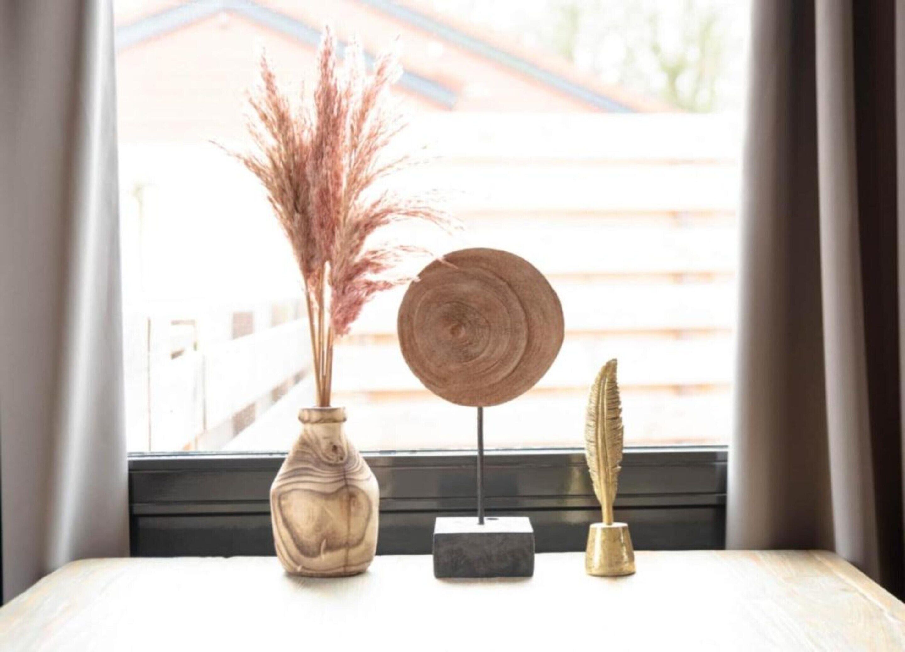 A window view with a wooden table displaying a vase with pampas grass, a wooden round plate, and a feather.