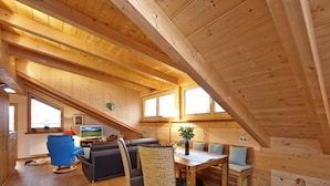 A spacious loft-style room with wooden beams, a sofa, a dining table, and a view of the outdoors. - Holiday Apartment Zugspitzblick-riedel (Garmisch-Partenkirchen)