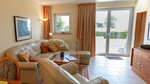 A living room with a sofa, armchairs, a coffee table, and a TV. There is a window with curtains, a radiator, and a door leading to an outdoor area. - Kaiserhof \"lake View\" (Kellenhusen)