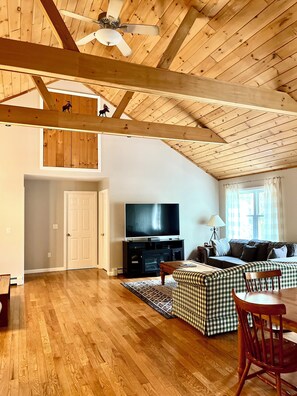 A spacious living room with a high ceiling, wooden beams, a large flat-screen TV, a sofa, and a dining area with a table and chairs. - Summer Getaway in White Mountain with Lake and Beach Access. Hiking and Swimming (Conway)