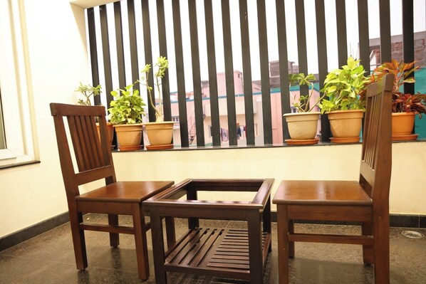 A balcony with two wooden chairs and a small table, potted plants, and a view of buildings through vertical blinds.