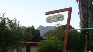 Ein Schild mit der Aufschrift 'GARDEN' in ländlicher Umgebung, mit einem Haus, Bäumen und Bergen im Hintergrund.