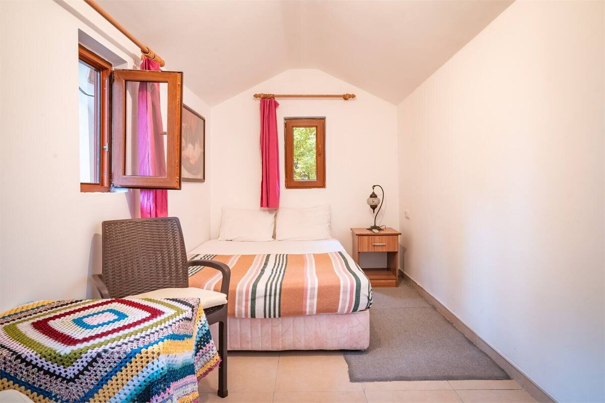 Un dormitorio pequeño y acogedor con una cama individual, una silla, una manta colorida y una mesita de noche con una lámpara.