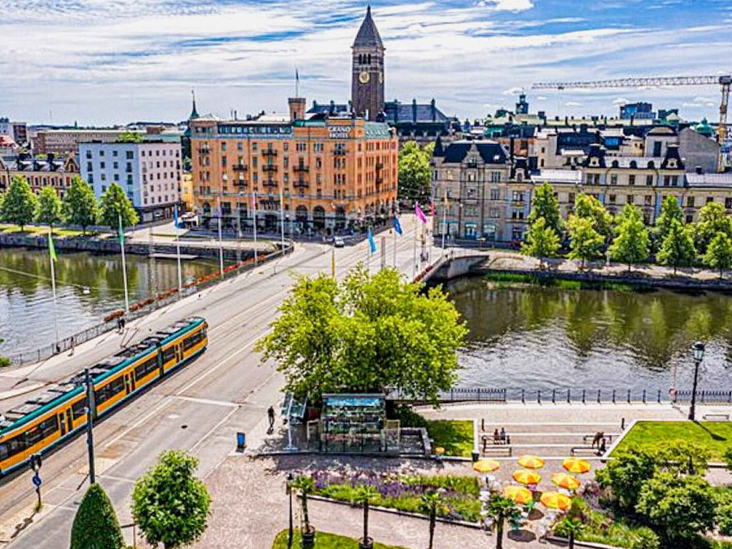 A riverside view of a city with a prominent clock tower, a tram line, and a riverside promenade with umbrellas.