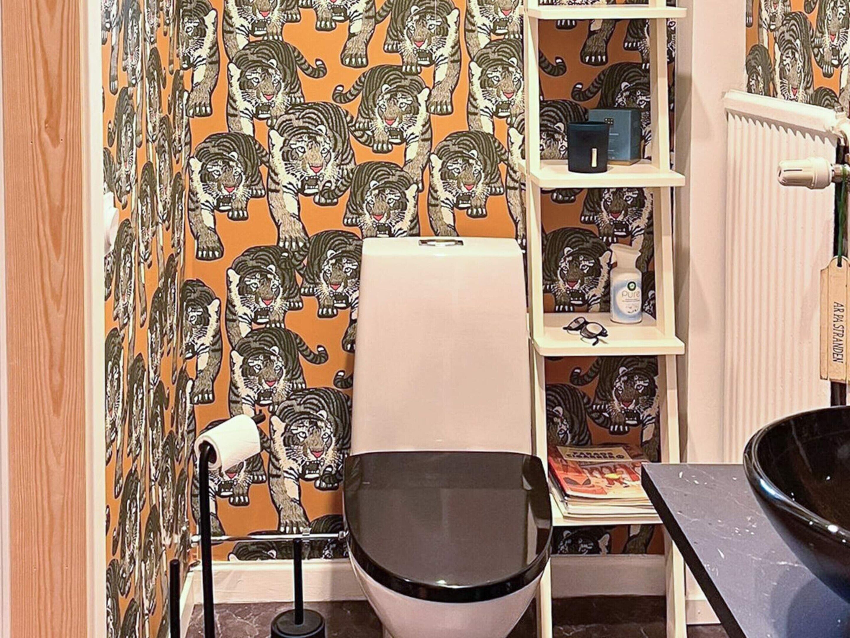 Un bany amb un vàter, una estantària amb articles de tocador i un paper pintat amb motius.