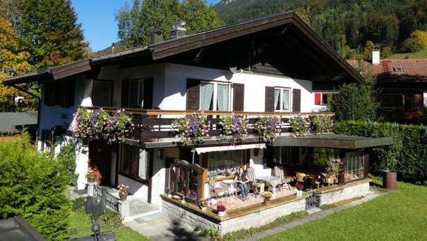 Un chalet de dos plantas con balcón, rodeado de exuberante vegetación y montañas al fondo.