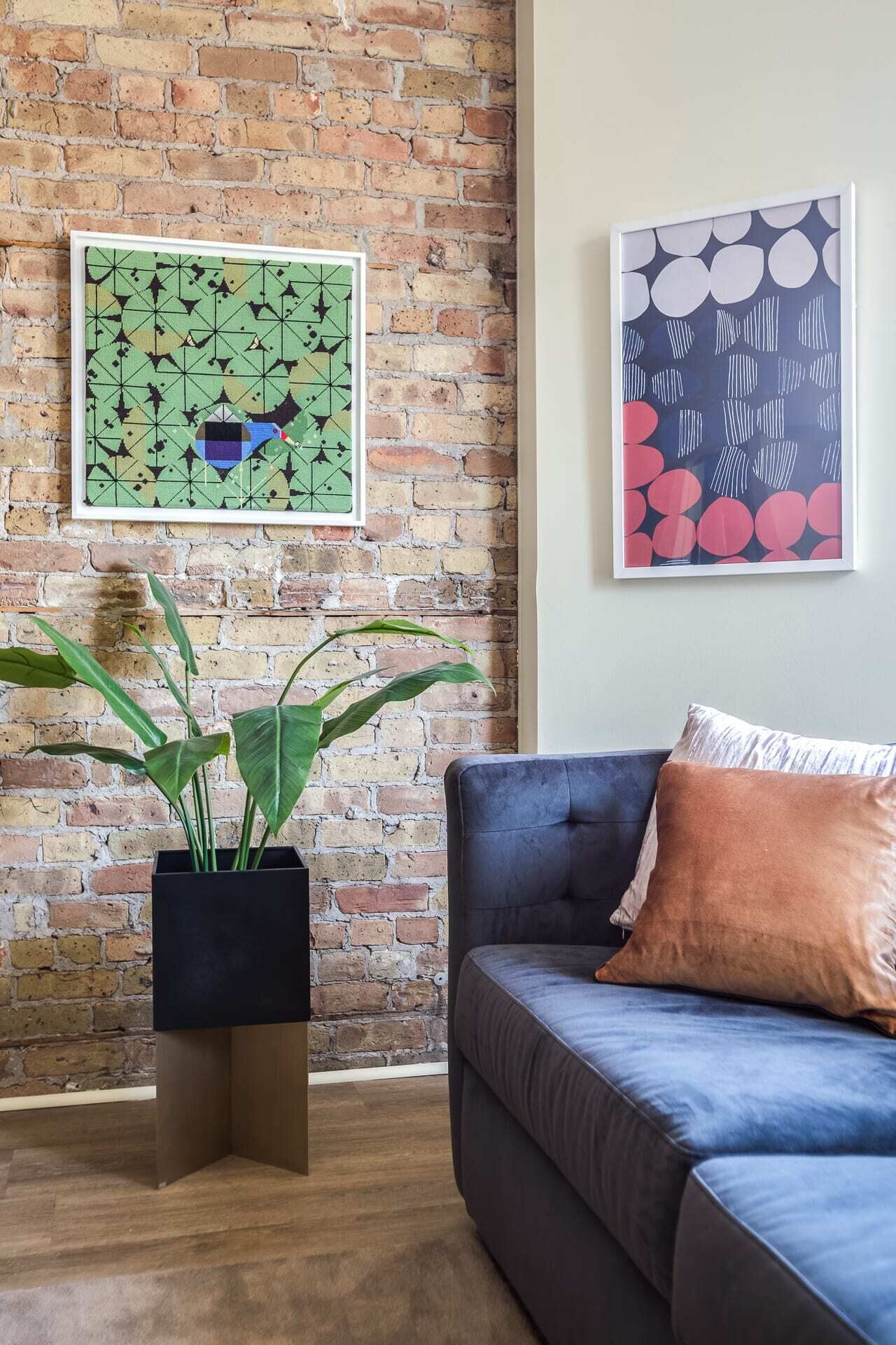 Ein Wohnzimmer mit Backsteinwand, einem blauen Sofa mit braunem Kissen und zwei gerahmten Kunstwerken.
