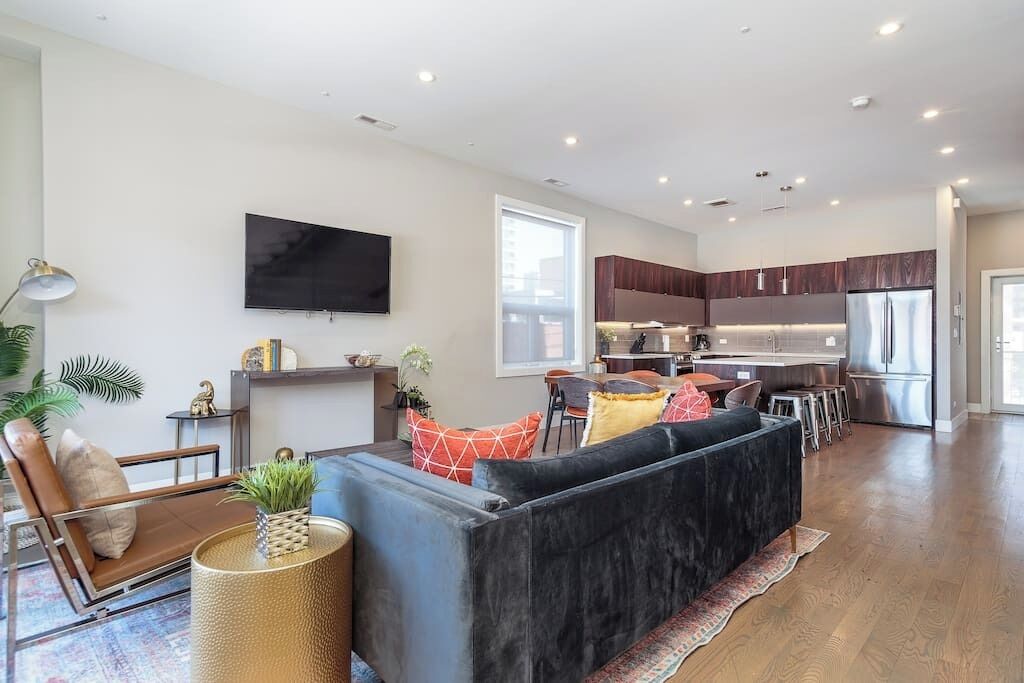 A modern living room with a black sofa, a coffee table, and a TV mounted on the wall. There is a kitchen area with wooden cabinets and a stainless steel refrigerator.