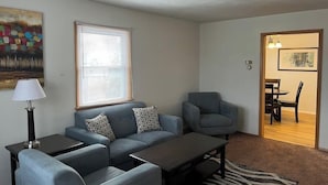 A modern living room with a grey sofa, matching armchairs, a coffee table, and a lamp. There is a painting on the wall and a window with blinds. - Close to Downtown - 2 queen beds (Springfield)