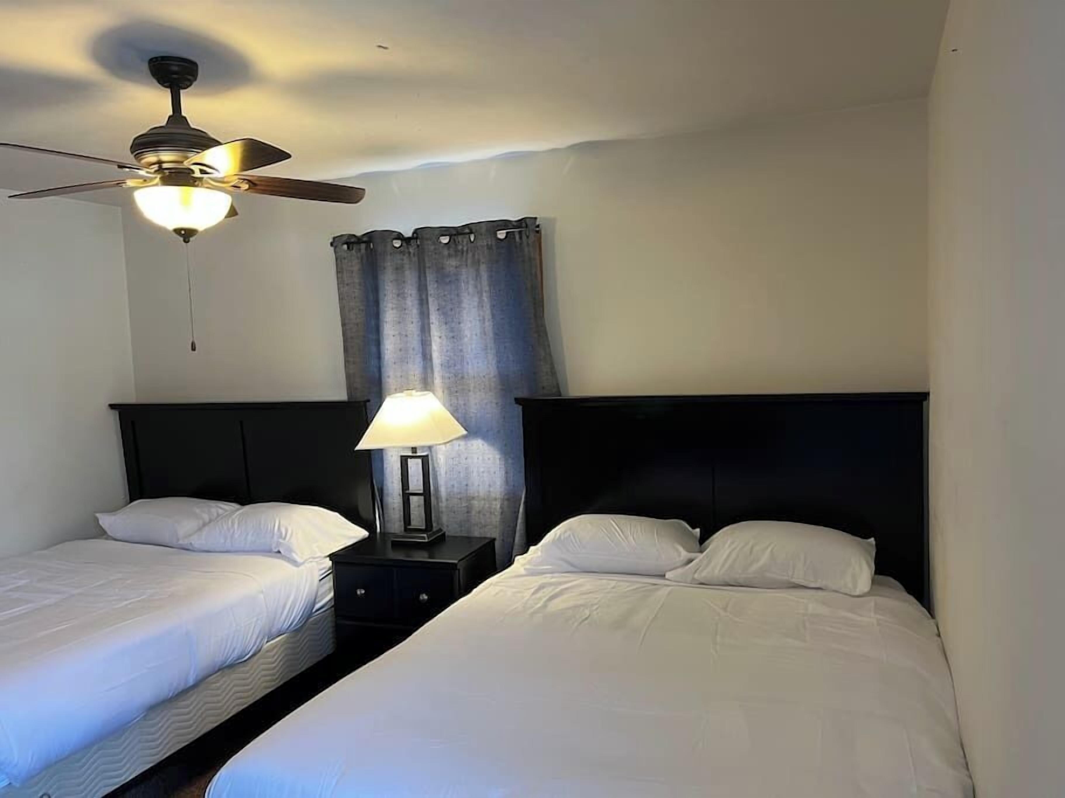 Habitación de hotel con dos camas individuales, un ventilador de techo, una mesita de noche con lámpara y una cortina.