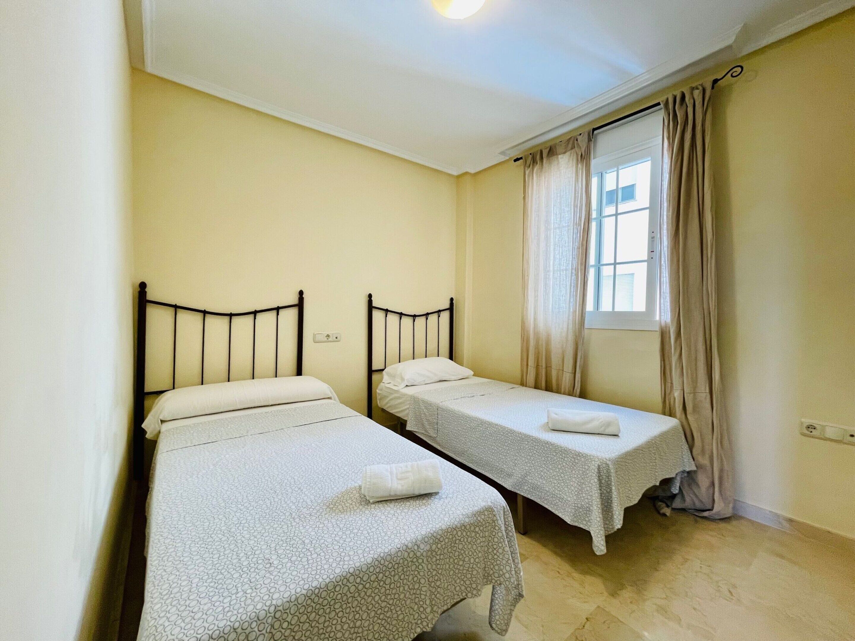 A small, clean room with two single beds, a window with curtains, and a light switch on the wall.