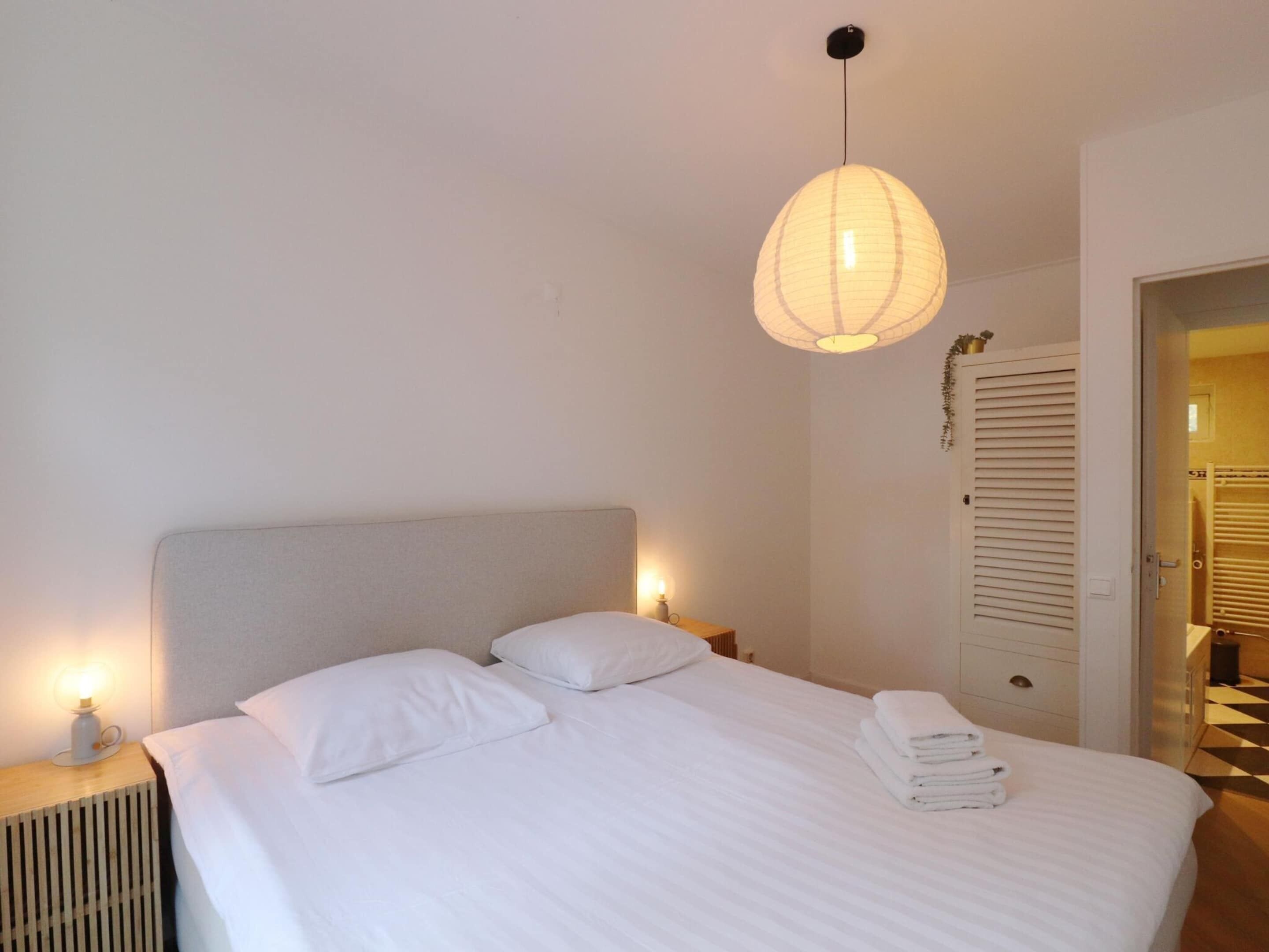 A neatly made bed with white linens, a bedside lamp, and a hanging light fixture.